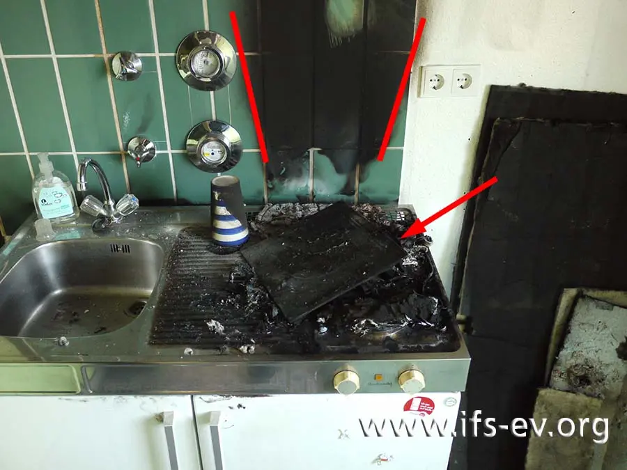 Vom Induktionskochfeld sind nur noch Reste vorhanden (Pfeil). An der Wand zeichnet sich ein Brandtrichter ab (Markierung).