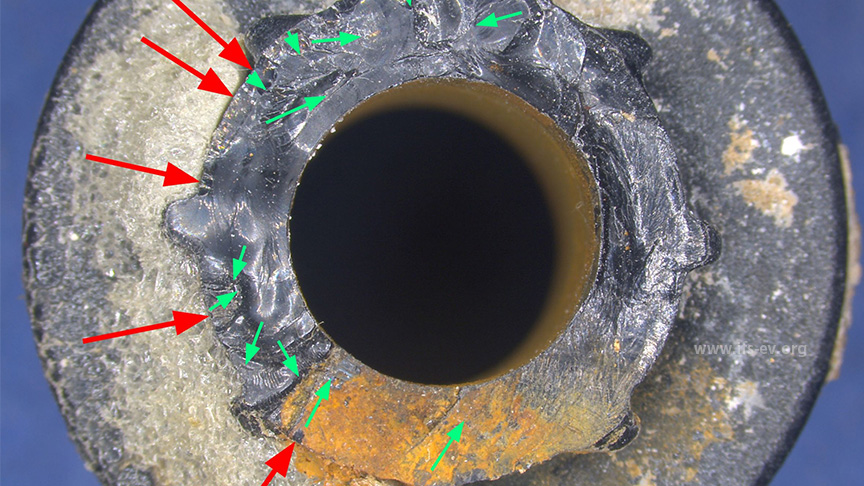 Mikroskopische Ansicht der Bruchfläche des abgebrochenen Teilstücks: Zahlreiche Rissausgangspunkte (exemplarisch rote Pfeile) und Rissausbreitungsrichtungen (exemplarisch grüne Pfeile) 